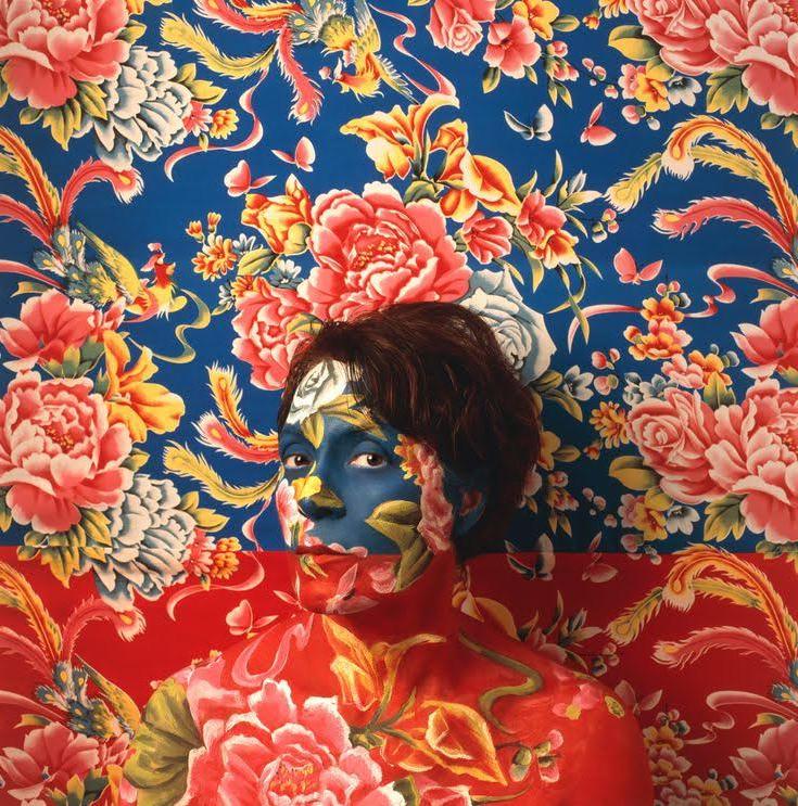 Self Portrait of Artist wearing blue and red floral make-up as camouflage with wallpaper behind her